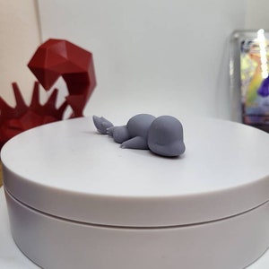 May include: A small, gray, 3D printed figurine of a Pokemon, possibly a Snorlax, lying on its side on a white circular base.