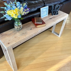 May include: A whitewashed wood console table with a distressed finish. The table has a rectangular top and a single open shelf. The table is decorated with a vase of flowers, two books, and a framed sign that says "Mostly Happy, Always Dreaming".