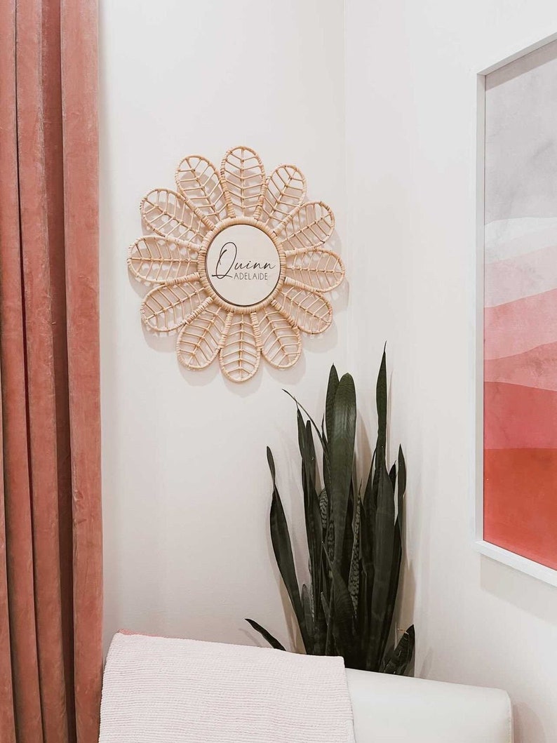 Puede incluir: Un espejo en forma de margarita hecho de rat&aacute;n tejido, con una secci&oacute;n central circular. El centro del espejo tiene el nombre "Quinn" y la palabra "Adelaide" en escritura negra. El espejo est&aacute; colgado en una pared blanca, con una cortina rosa y una obra de arte.