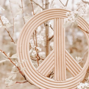 K&ouml;nnte beinhalten: Ein Wanddekor aus Naturholz in Form eines Friedenszeichens mit geflochtenem Design. Das Friedenszeichen h&auml;ngt an einem Baumzweig mit wei&szlig;en Bl&uuml;ten.