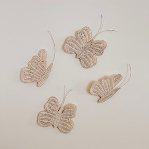 May include: Four woven butterfly wall decorations in a light beige color. Each butterfly has detailed wing patterns and thin, curved antennae. The butterflies are arranged on a plain, off-white background.