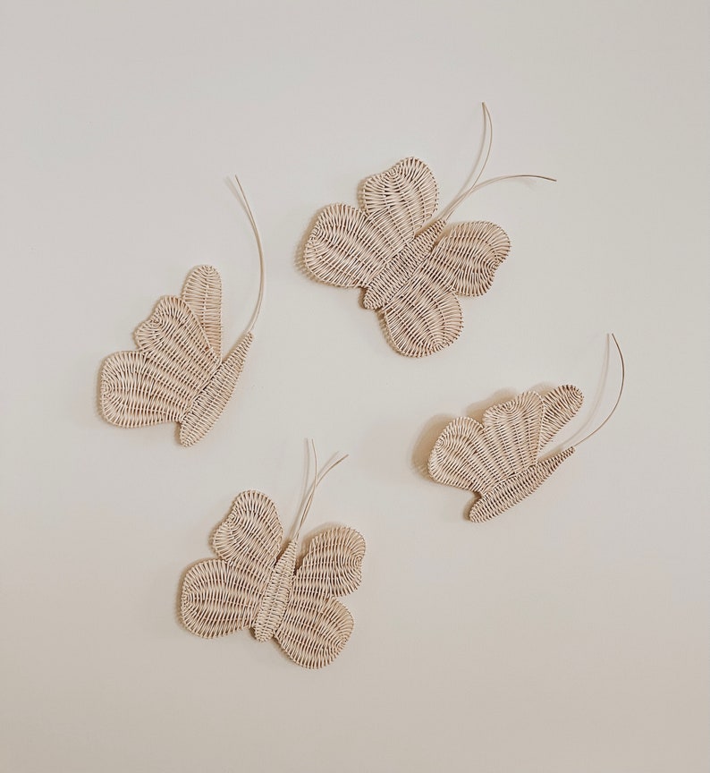 May include: Four woven butterfly wall decorations in a light beige color. Each butterfly has detailed wing patterns and thin, curved antennae. The butterflies are arranged on a plain, off-white background.