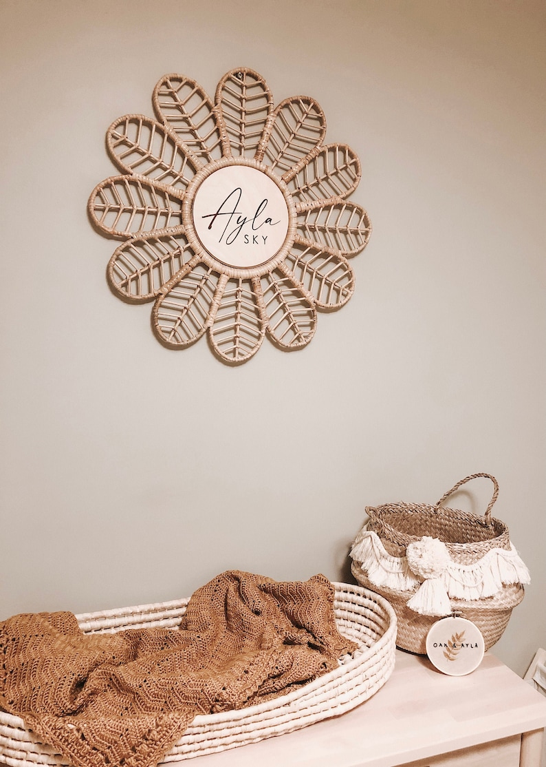 Puede incluir: Decoraci&oacute;n de pared en forma de flor de rat&aacute;n tejido con el nombre "Ayla Sky" en escritura negra. Debajo, una cesta tejida contiene una manta marr&oacute;n. Tambi&eacute;n hay una segunda cesta con borlas y un peque&ntilde;o aro bordado.