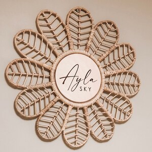Puede incluir: Decoraci&oacute;n de pared de rat&aacute;n tejido en forma de flor. El c&iacute;rculo central es de madera clara y presenta el nombre "Ayla SKY" en escritura negra. Los p&eacute;talos de la flor est&aacute;n intrincadamente tejidos, creando una est&eacute;tica natural y bohemia.