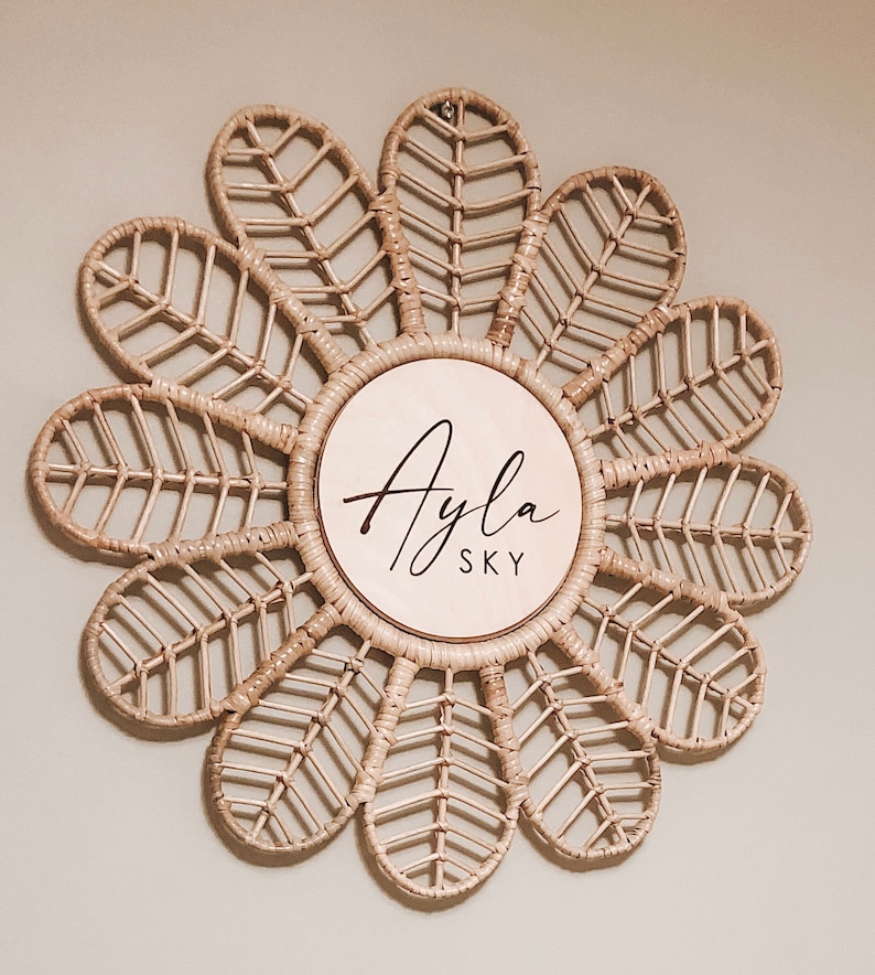 Puede incluir: Decoraci&oacute;n de pared de rat&aacute;n tejido en forma de flor. El c&iacute;rculo central es de madera clara y presenta el nombre "Ayla SKY" en escritura negra. Los p&eacute;talos de la flor est&aacute;n intrincadamente tejidos, creando una est&eacute;tica natural y bohemia.