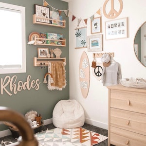 K&ouml;nnte beinhalten: Ein Kinderzimmer mit einer gr&uuml;nen Wand, einem wei&szlig;en Sitzsack, einem Surfboard aus Holz und einer Kommode mit drei Schubladen. Das Zimmer ist mit einem Friedenssymbol, einem Regenbogen und einem Holzschild mit der Aufschrift "Rhodes" dekoriert.