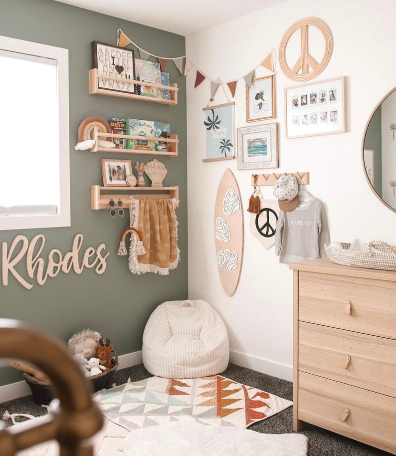 K&ouml;nnte beinhalten: Ein Kinderzimmer mit einer gr&uuml;nen Wand, einem wei&szlig;en Sitzsack, einem Surfboard aus Holz und einer Kommode mit drei Schubladen. Das Zimmer ist mit einem Friedenssymbol, einem Regenbogen und einem Holzschild mit der Aufschrift "Rhodes" dekoriert.