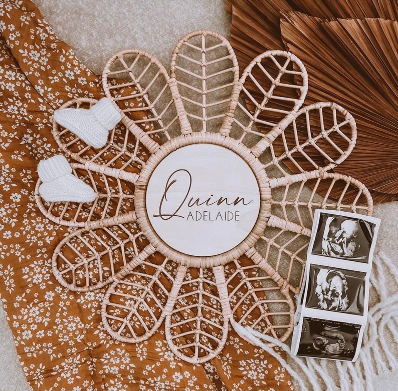 Puede incluir: Decoraci&oacute;n de pared en forma de flor de rat&aacute;n tejido con un centro de madera que muestra el nombre "Quinn Adelaide". Dos botines de beb&eacute; blancos e im&aacute;genes de ultrasonido se colocan cerca de la decoraci&oacute;n. El fondo incluye una manta con estampado floral.