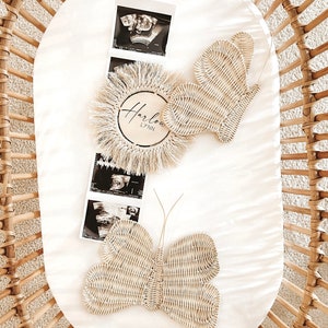 May include: A wicker crib with a white mattress pad holds a decorative arrangement. The arrangement includes two woven butterfly decorations, ultrasound images, and a wooden disc with the name "Harlow Lynn" in script. The butterflies and disc are light beige.
