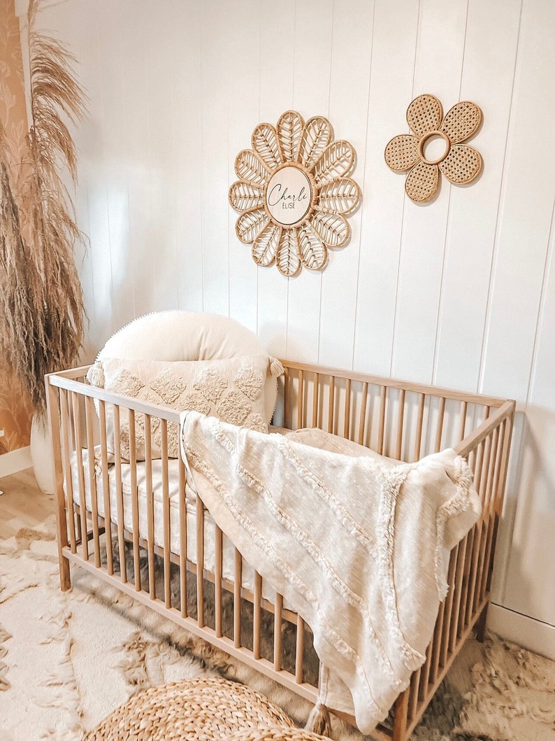 Puede incluir: Una cuna de madera clara con una manta y ropa de cama de color crema. Dos decoraciones de flores tejidas cuelgan de la pared blanca sobre la cuna. Una decoraci&oacute;n floral tiene el nombre "Charlie Elise" escrito.
