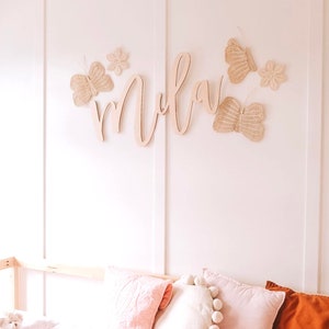 May include: A wall decoration featuring a wooden name and butterfly accents. The name is in a cursive font, and the butterflies are made of woven material. The decor is mounted on a white wall, with pink pillows and a teddy bear visible below.