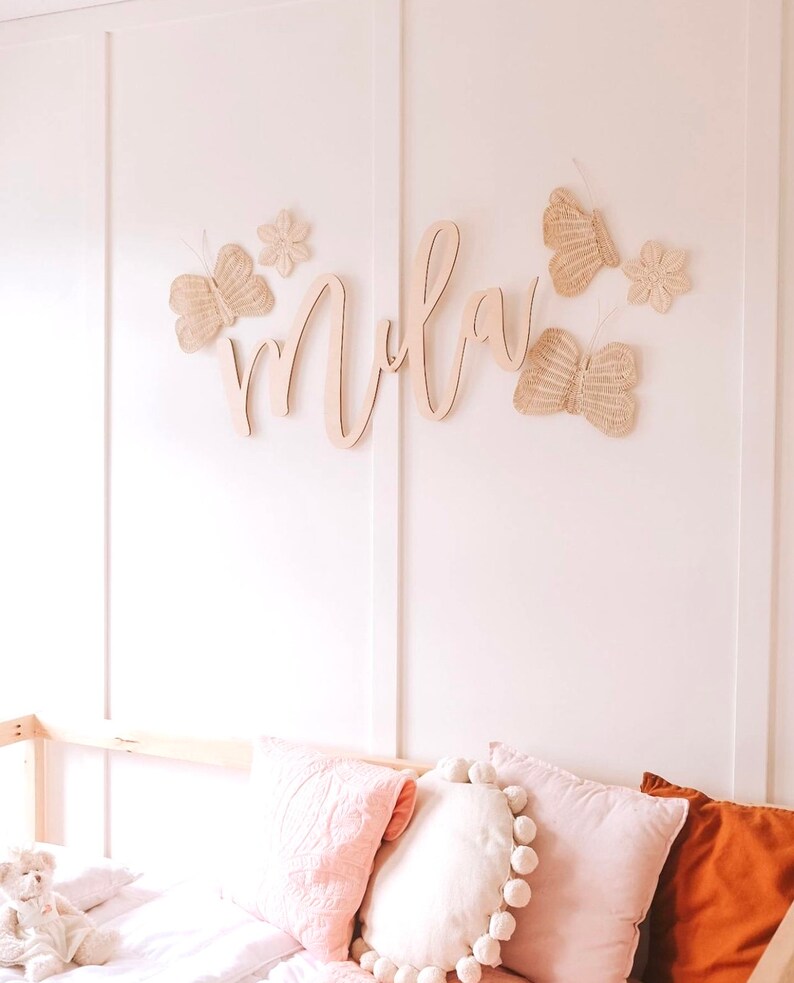 May include: A wall decoration featuring a wooden name and butterfly accents. The name is in a cursive font, and the butterflies are made of woven material. The decor is mounted on a white wall, with pink pillows and a teddy bear visible below.