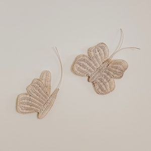 May include: Two decorative butterfly wall hangings. The butterflies are made of woven, off-white material. Each butterfly has a curved, wire-like antenna. The butterflies are mounted on a plain, off-white wall.