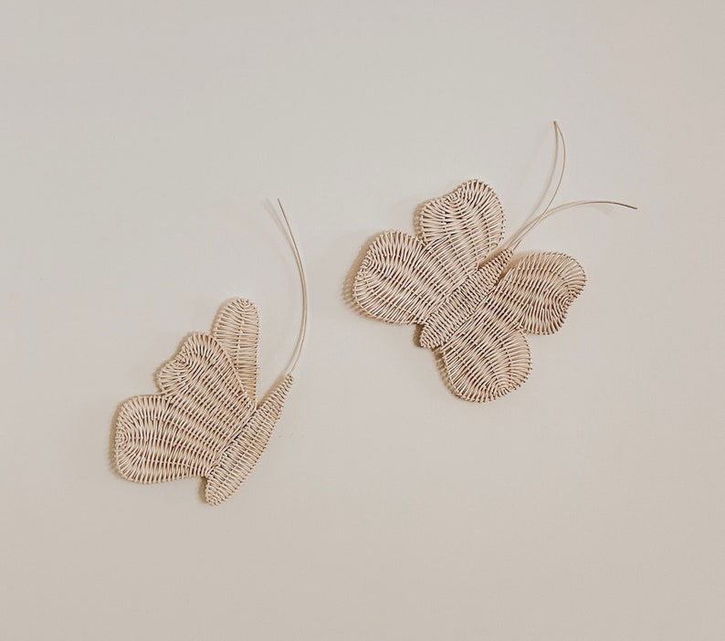 May include: Two decorative butterfly wall hangings. The butterflies are made of woven, off-white material. Each butterfly has a curved, wire-like antenna. The butterflies are mounted on a plain, off-white wall.