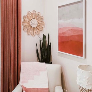Puede incluir: Un interior de sala de estar con un sill&oacute;n blanco y una manta estampada en rosa y blanco. Una obra de arte abstracta enmarcada y un espejo tejido en forma de flor con el texto "Quinn Adelaide" adornan la pared. Se ve una cortina rosa.