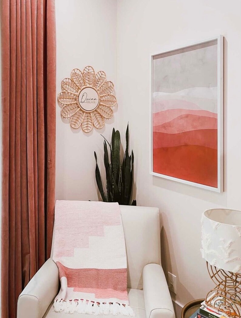 Puede incluir: Un interior de sala de estar con un sill&oacute;n blanco y una manta estampada en rosa y blanco. Una obra de arte abstracta enmarcada y un espejo tejido en forma de flor con el texto "Quinn Adelaide" adornan la pared. Se ve una cortina rosa.