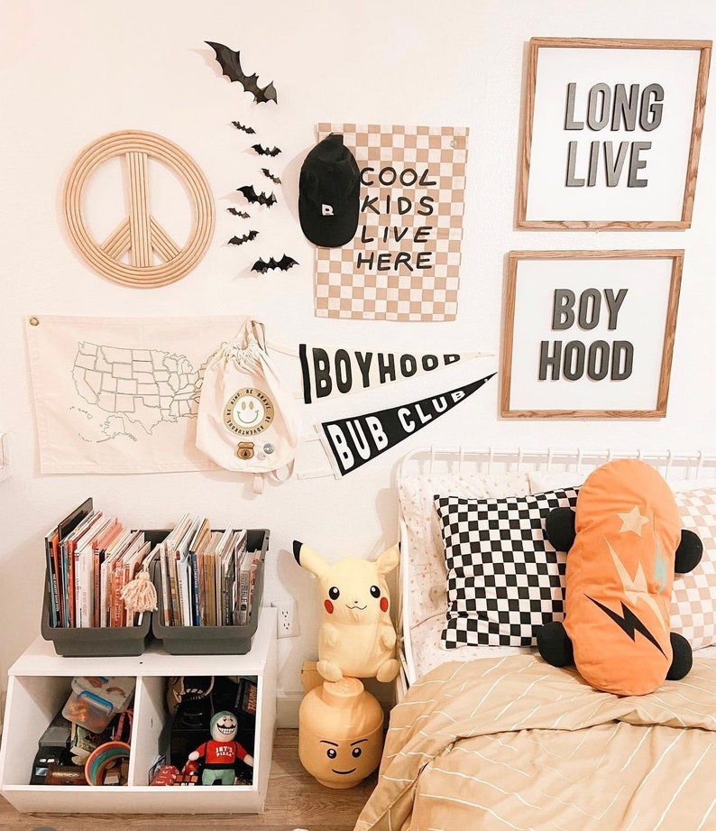 K&ouml;nnte beinhalten: Ein wei&szlig;es Bett mit einem beige-wei&szlig;en gestreiften Bettbezug und einem schwarz-wei&szlig; karierten Kissen. Auf dem Bett liegen zwei Kissen, eines ist orange mit einem Blitz-Design und das andere ist ein gelbes Pikachu-Pl&uuml;schtier. An der Wand hinter dem Bett h&auml;ngen mehrere gerahmte Schilder, darunter eines mit der Aufschrift "Long Live" und ein anderes mit der Aufschrift "Boyhood".
