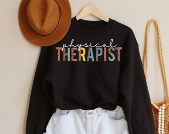 May include: Black sweatshirt with the text "physical therapist" in colorful letters. The sweatshirt is worn with denim shorts and a straw bag.