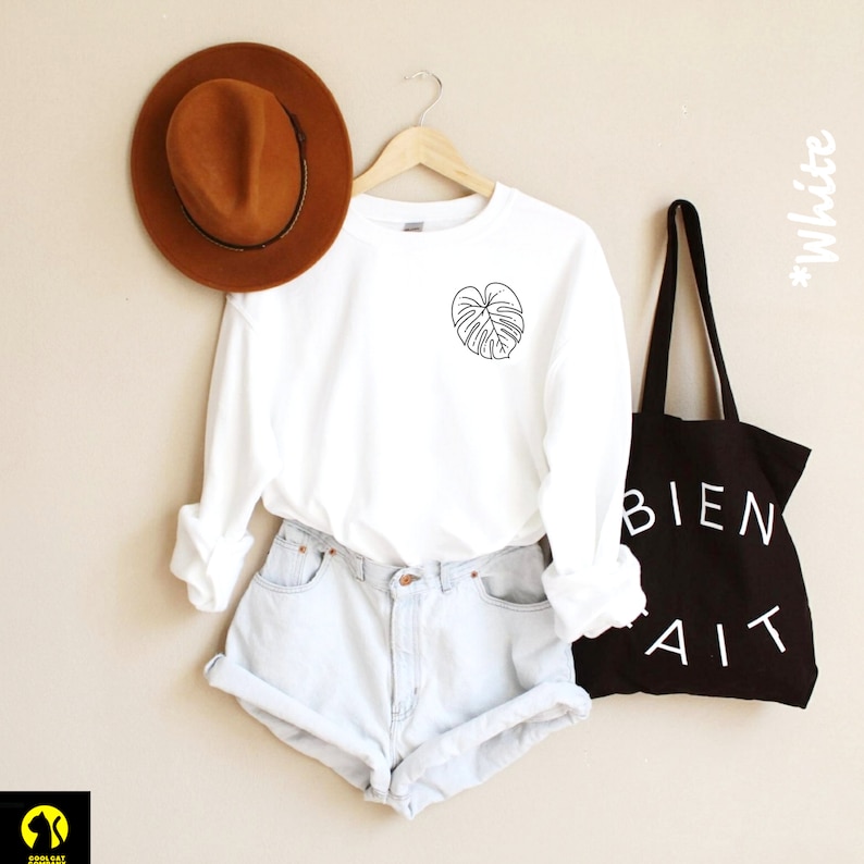 May include: White long-sleeved shirt with a black outline of a leaf on the left chest. The shirt is worn with light blue denim shorts and a brown fedora. A black tote bag with the text "BIEN FAIT" in white is on the right side of the image.