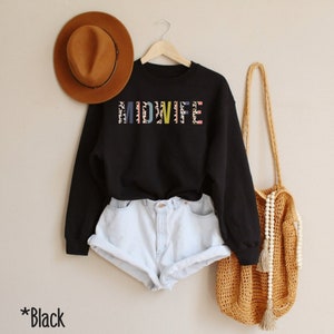 Pode incluir: Camisola preta com a palavra "MIDWIFE" impressa em letras coloridas com um padr&atilde;o de leopardo. A camisola est&aacute; sendo modelada com um par de cal&ccedil;&otilde;es de ganga azul claro e uma bolsa de palha.