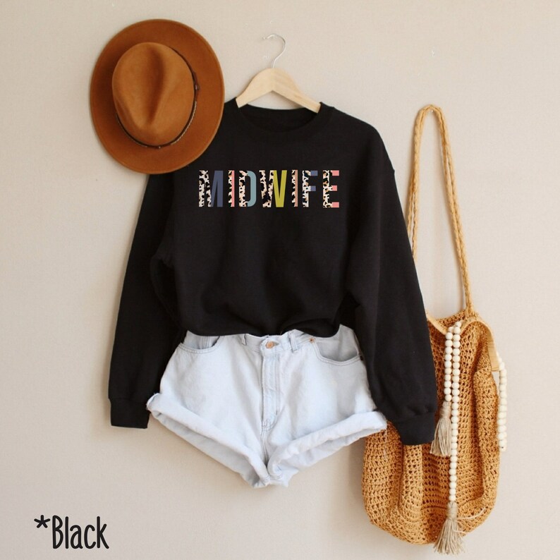 Pode incluir: Camisola preta com a palavra "MIDWIFE" impressa em letras coloridas com um padr&atilde;o de leopardo. A camisola est&aacute; sendo modelada com um par de cal&ccedil;&otilde;es de ganga azul claro e uma bolsa de palha.