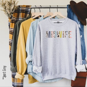 Pode incluir: Uma camisa de moletom cinza com a palavra "MIDWIFE" impressa em letras coloridas com um padr&atilde;o de leopardo. A camisa de moletom est&aacute; pendurada em um cabide com outras roupas.