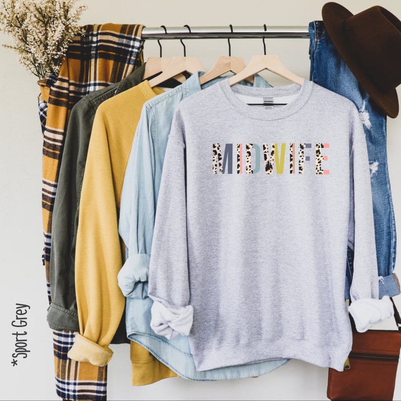 Pode incluir: Uma camisa de moletom cinza com a palavra "MIDWIFE" impressa em letras coloridas com um padr&atilde;o de leopardo. A camisa de moletom est&aacute; pendurada em um cabide com outras roupas.