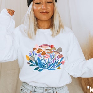 May include: White long-sleeved sweatshirt with a colourful mushroom and floral graphic print. The text "*White" is visible in the bottom right corner of the image.
