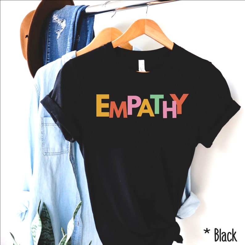 May include: Black t-shirt with the word "EMPATHY" in colorful block letters. The shirt is hanging on a wooden hanger with other clothing items in the background. The word "Black" is written in the bottom right corner.