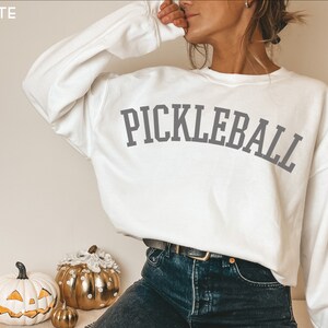 May include: White sweatshirt with the text "PICKLEBALL" in grey lettering. The sweatshirt is being worn by a person with long brown hair and is paired with blue jeans.