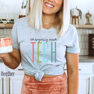 Puede incluir: Una camiseta gris con el texto "emergency room TECH" en letras de colores. La camiseta est&aacute; anudada a la cintura y la modelo sostiene una taza rosa y blanca.