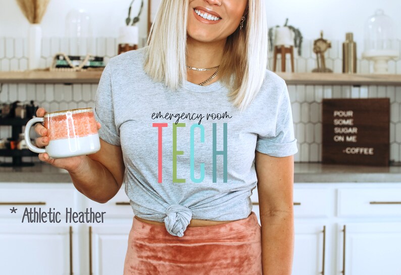 Puede incluir: Una camiseta gris con el texto "emergency room TECH" en letras de colores. La camiseta est&aacute; anudada a la cintura y la modelo sostiene una taza rosa y blanca.