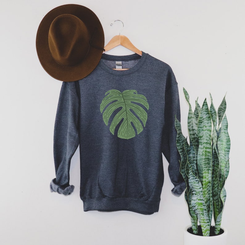Peut inclure: Un sweat-shirt gris fonc&eacute; avec un motif de feuille de monstera verte sur le devant. Le sweat-shirt est suspendu &agrave; un cintre en bois.