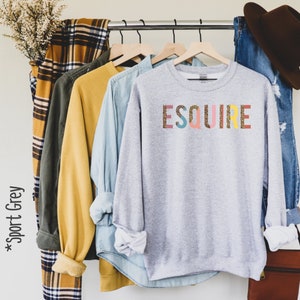 Peut inclure: Un sweat-shirt gris avec le mot "ESQUIRE" imprim&eacute; en lettres color&eacute;es avec un motif de paillettes dor&eacute;es. Les lettres sont rose, jaune, bleu, vert et rouge.