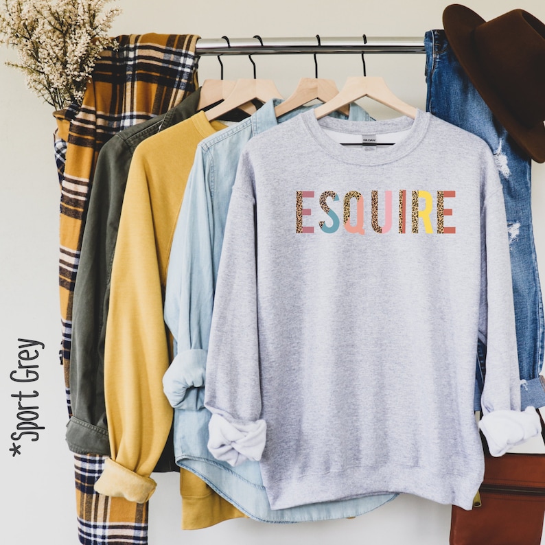 Peut inclure: Un sweat-shirt gris avec le mot "ESQUIRE" imprim&eacute; en lettres color&eacute;es avec un motif de paillettes dor&eacute;es. Les lettres sont rose, jaune, bleu, vert et rouge.