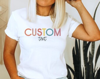 Könnte beinhalten: Weißes T-Shirt mit dem Text "CUSTOM text" in bunten Buchstaben. Die Buchstaben sind in einem Kreis umrandet, wobei jeder Kreis eine andere Farbe hat.