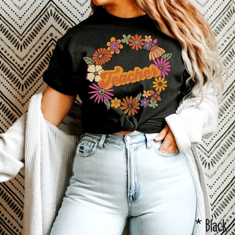K&ouml;nnte beinhalten: Ein schwarzes T-Shirt mit einem floralen Kranzdesign und dem Wort "Teacher" in einer Retro-Schrift. Die Blumen sind in Orange-, Gelb-, Rosa- und Gr&uuml;nt&ouml;nen gehalten.