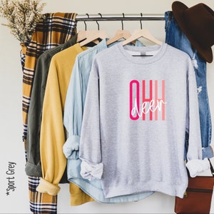 May include: Gray sweatshirt with the text "OHH deer" in pink and white lettering. The sweatshirt is hanging on a rack with other clothing.