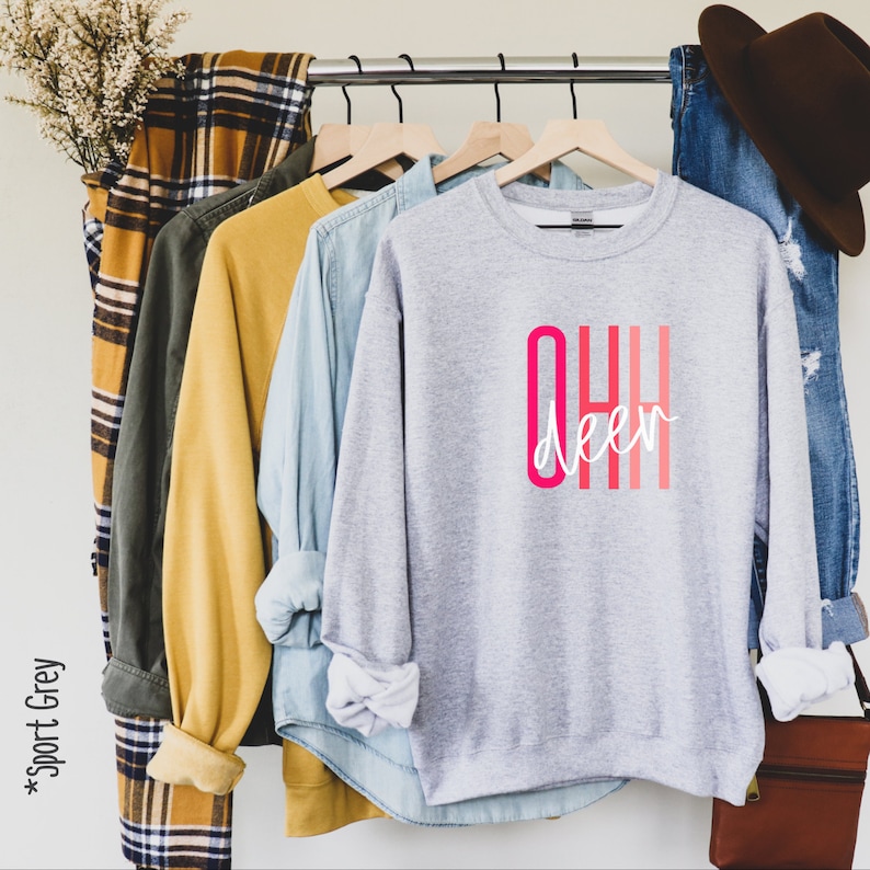May include: Gray sweatshirt with the text "OHH deer" in pink and white lettering. The sweatshirt is hanging on a rack with other clothing.