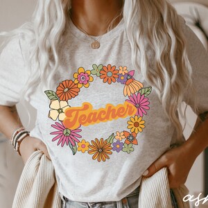 K&ouml;nnte beinhalten: Ein hellgraues T-Shirt mit einem Blumengirlanden-Design und dem Wort "Teacher" in einer Retro-Schrift. Die Blumen sind in den Farben Rosa, Gelb, Orange und Gr&uuml;n gehalten.