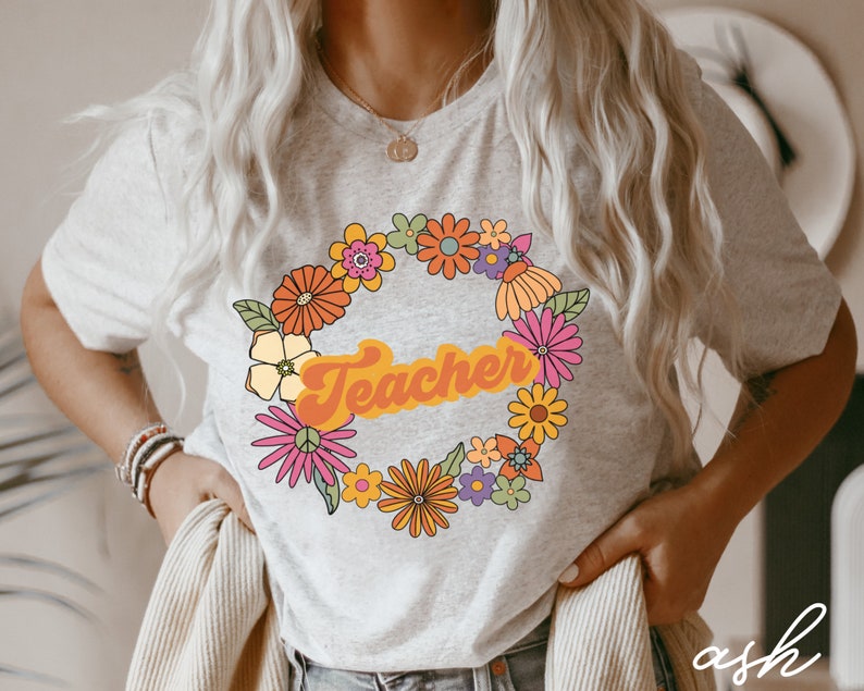 K&ouml;nnte beinhalten: Ein hellgraues T-Shirt mit einem Blumengirlanden-Design und dem Wort "Teacher" in einer Retro-Schrift. Die Blumen sind in den Farben Rosa, Gelb, Orange und Gr&uuml;n gehalten.