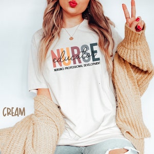 May include: A white t-shirt with the text "NURSE educator NURSING PROFESSIONAL DEVELOPMENT" in rainbow colors. The text is in a stylized font. The shirt is being worn by a person with long blonde hair and a tan cardigan.