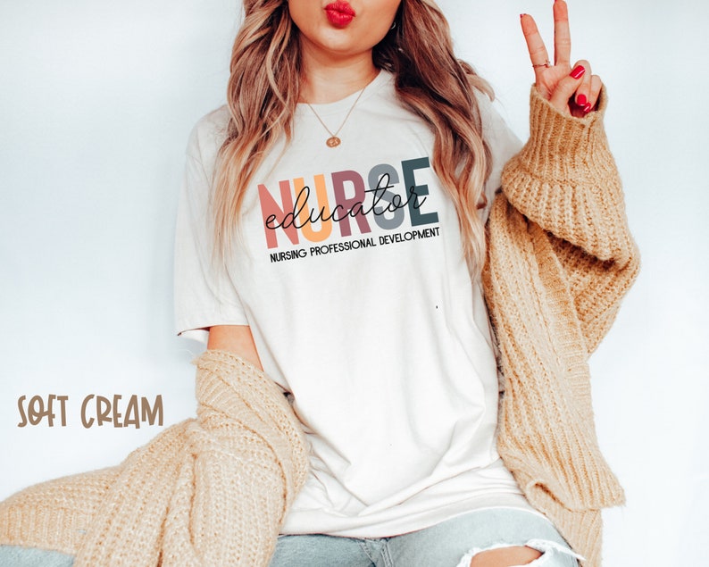 May include: A white t-shirt with the text "NURSE educator NURSING PROFESSIONAL DEVELOPMENT" in rainbow colors. The text is in a stylized font. The shirt is being worn by a person with long blonde hair and a tan cardigan.