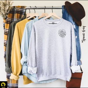 May include: A gray sweatshirt with a black outline of a monstera leaf on the left chest. The sweatshirt is hanging on a rack with other clothing.