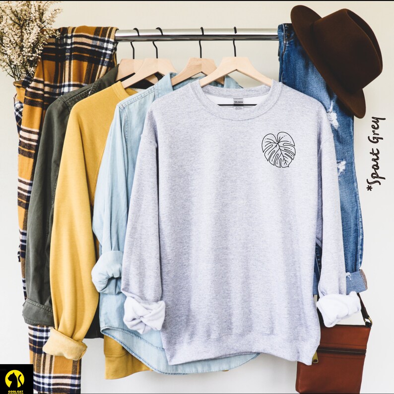 May include: A gray sweatshirt with a black outline of a monstera leaf on the left chest. The sweatshirt is hanging on a rack with other clothing.