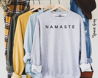 Może przedstawiać: Szara bluza z nadrukiem "NAMASTE" w kolorze czarnym na przodzie. Bluza wisi na wieszaku z innymi ubraniami.