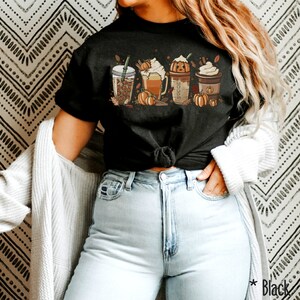 May include: A black t-shirt with a graphic of four fall-themed drinks. The drinks include a pumpkin spice latte, a caramel macchiato, a pumpkin spice cold brew, and a peppermint mocha. The graphic is surrounded by fall leaves and pumpkins.
