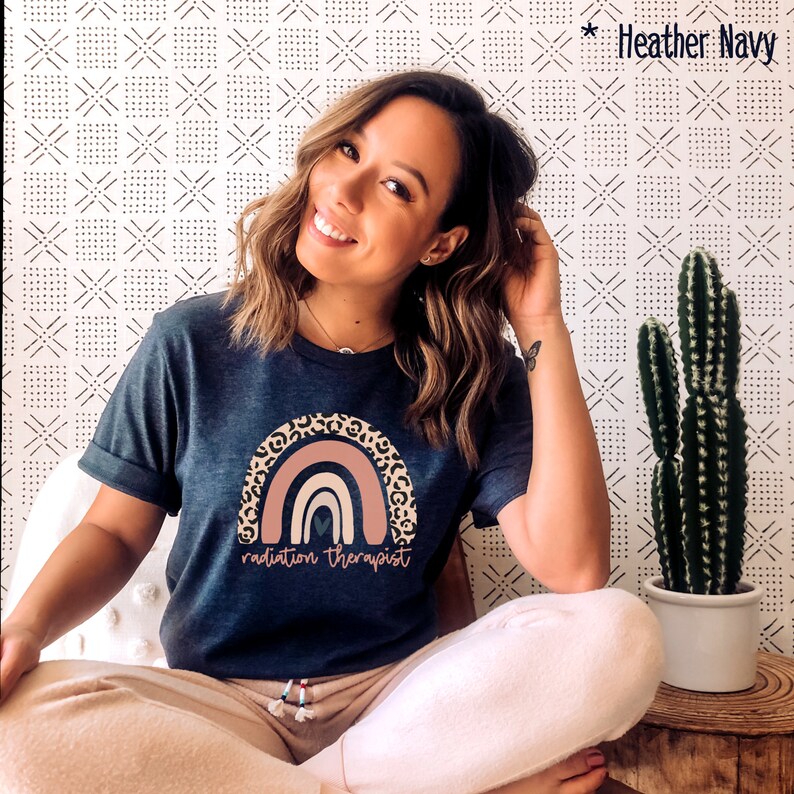 Puede incluir: Una camiseta azul marino con un gr&aacute;fico de arco&iacute;ris y el texto "radiation therapist". El arco&iacute;ris est&aacute; hecho de rayas rosas, naranjas y marrones con un borde de estampado de leopardo.