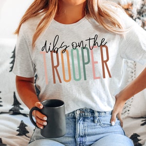 Peut inclure: Un t-shirt blanc avec le texte "dibs on the TROOPER" en lettres colorées. Les lettres sont dans des tons de brun, rose, vert et bleu.