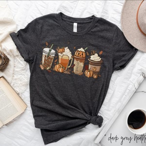 May include: A dark grey heather t-shirt with a graphic of four fall-themed drinks. The drinks include a pumpkin spice latte, a pumpkin-shaped drink, a caramel macchiato, and a regular coffee. Each drink is topped with whipped cream and a sprinkle of cinnamon. The graphic also includes fall leaves and pumpkins.